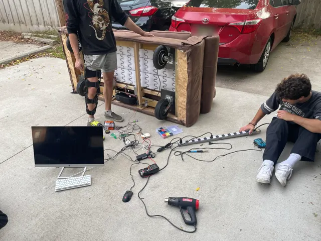 The couch tipped on its back in a driveway with wheels fitted while friends wire it up, laptop and tools on the ground